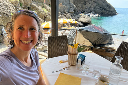 Woman smiling at a seaside restaurant in San Fruttuoso, with yellow umbrellas and turquoise water in the background.