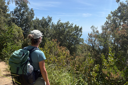 Hiker with a green backpack standing on a coastal trail surrounded by trees, overlooking the Ligurian Sea near Portovenere.