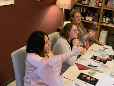 Three women and a man sitting at a table for a wine tasting. Two women are holding out and examining their wine glasses.