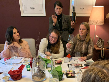 Women sitting at a table with food and wine glasses. Sommelier in the background explainining wine.
