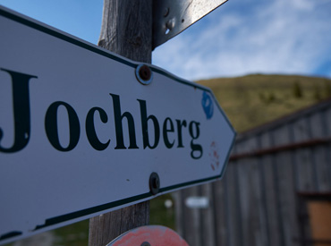 Trail sign pointing to Jochberg in the Bavarian Alps with wooden hut and grassy slope in the background – captured during a guided mountain hike near Walchensee.
