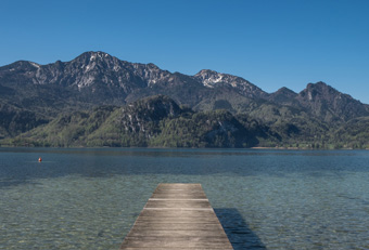 View over Lake Kochel in the Bavarian Alps with calm turquoise water, forested shore and village houses under a clear blue sky – typical scenery of the Four Elements guided nature hikes.