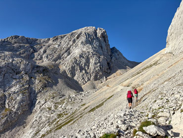 Hikers walking along a mountain trail beneath towering limestone peaks in Slovenia's Julian Alps