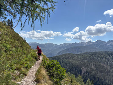 Two hikers walking along a narrow mountain trail above a forested valley in Slovenia's Julian Alps