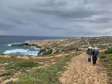 Two walkers walking uphill on sandy footpath with ocean and cliffs in background